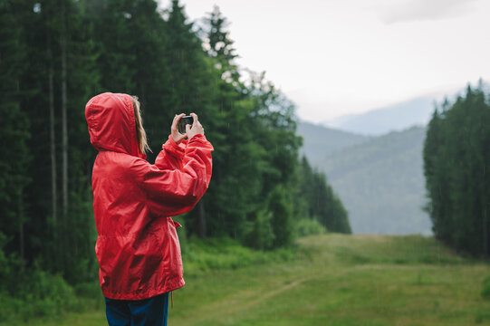 Hiker woman in red raincoat stands on background of beautiful mountain landscape in rainy weather and takes photo on smartphone. Girl taking photo of landscape in mountains during rain in hike.