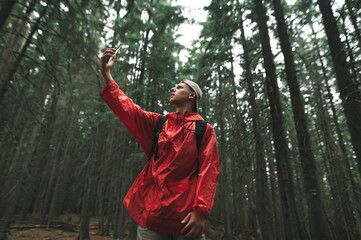 Man in a red raincoat stands in a thick coniferous forest on a hike and catches a phone call on a smartphone with his arm raised up.Hiker in the mountains looking for a mobile network on a smartphone.