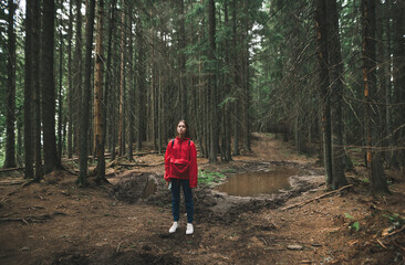 Obraz premium Hiker woman in a red raincoat stands in the forest on a mountain path on a puddle background with a swamp and looks camera.Hiking through the mountains through the forest. Hiker girl on a hiking trip.