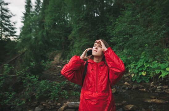 Concerned girl tourist stands in the forest in a red raincoat, calls on phone and looks away with a worried face in a mountain hike against a backdrop of a mountain stream and coniferous trees.