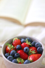 Bowl with blueberries and strawberries and open book on a bed. Selective focus. 