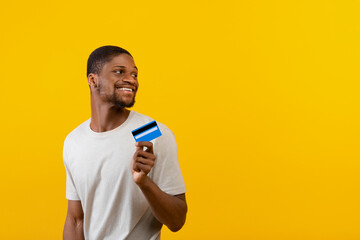 Excited african american man holding credit card and looking at empty space on yellow studio background
