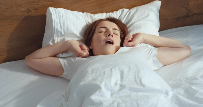 Hidden camera view of the young ginger woman waking up after sleeping in her bed and stretching in the morning. Woman yawns and smiling pleasure. Calm and peaceful mood concept. Stock video