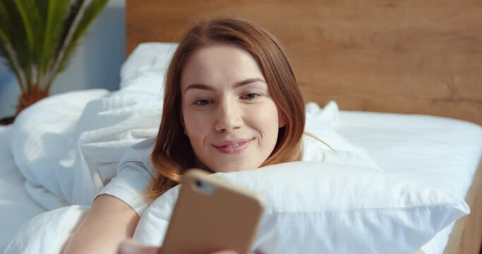 Close up view of the happy fresh woman with ginger hair making selfie in bed while waking up alone. Smiling young lady awake after healthy sleep and laying in cozy comfortable bedroom interior