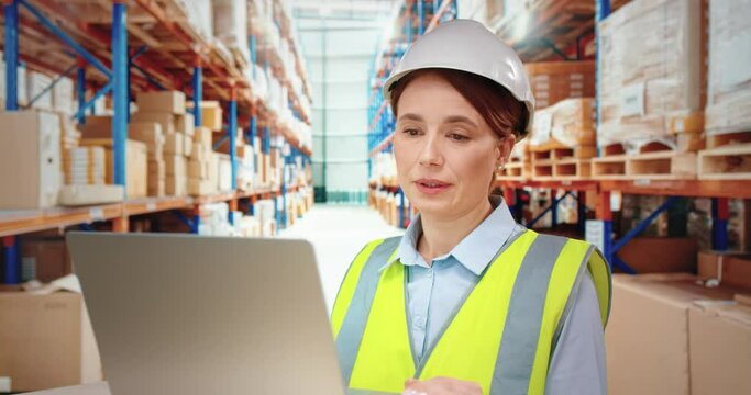 Beautiful female warehouse worker in white helmet talks on video call using laptop while standing on background of rows with cargo.