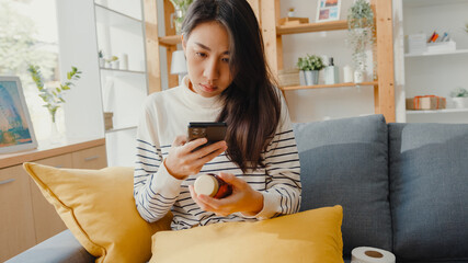 Sick young Asian woman hold medicine sit on couch take photo sent to doctor at home. Girl take medicine after doctor order, quarantine at house, Coronavirus social distancing quarantine concept.