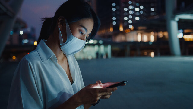 Young Asia businesswoman in fashion clothes wearing face mask using smart phone typing text message while stand outdoor in urban city at night. Social distancing to prevent spread of COVID-19 concept.