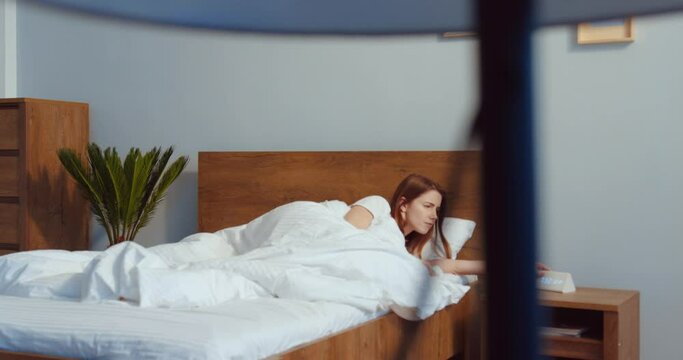 Motion camera view of the ginger caucasian woman looking attentively at the alarm clock and hiding under the white blanket because don't want to get up. Lazy morning concept