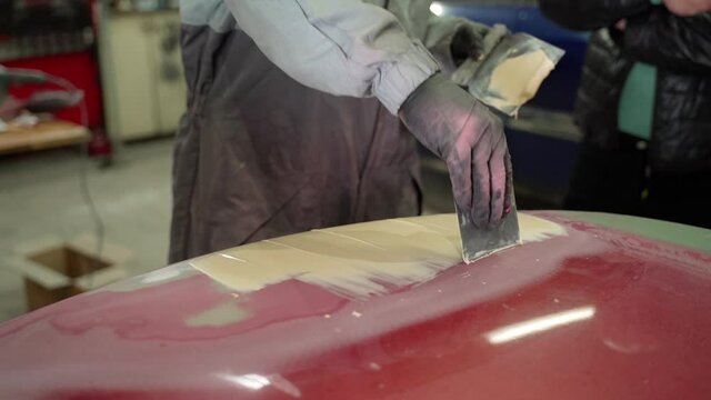 Man auto mechanic uses a spatula to put putty on the damaged part of the car's hood. Close-up shot of automobile refurbishment work