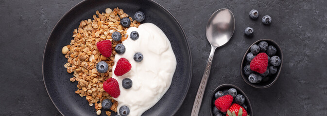 Horizontal banner with homemade granola, yogurt and fresh berries in black ceramic plate on dark stone background. Healthy vegan breakfast
