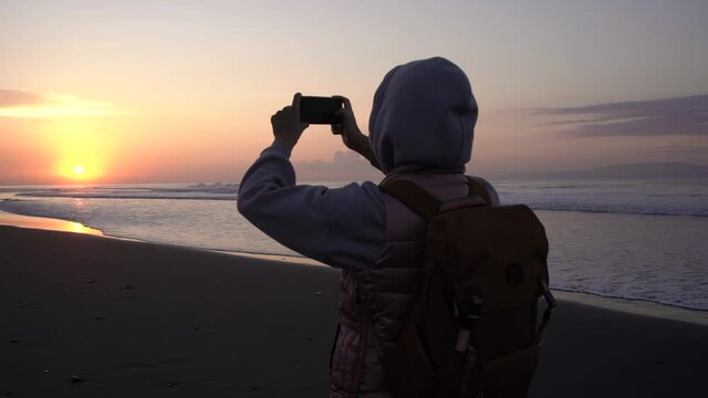 Rear view young woman traveler with backpack hood on head stands by the sea and enjoys the sunrise takes photos on your smartphone.