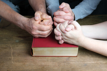 close-up of the hands of two men and a woman folded in a prayer gesture on a thick family bible in...