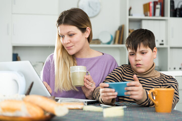 Portrait of young mother and son with tea using gadgets at table indoors