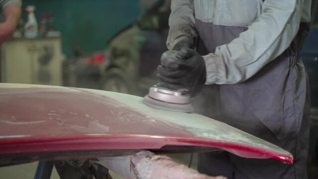 A man auto mechanic uses a grinder to grind a putty on a damaged car body part. Repair work to restore the paintwork in the garage by a professional specialist concept. Close-up shot