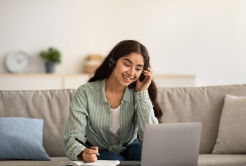 Happy Indian businesswoman in headphones having online business meeting on laptop, writing down info at home