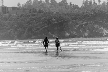 Para surfer&oacute;w, dziewczyna i chłopak idący z deskami surfingowymi po plaży na tle oceanu i klifu.