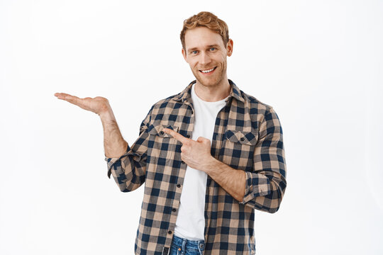 Image of smiling redhead man pointing at his open hand, display an item, recommending product on his palm, showing object, standing against white background