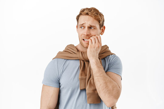 Image of anxious red head man biting finger nails and looking left with scared worried face, staring at something with anxiety, standing nervous against white background
