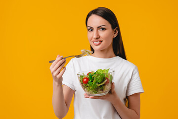 Woman caucasian latin eating salad from salad bowl in white T-shirt isolated on yellow background....
