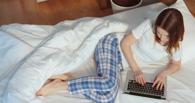 Horizontal view of the calm caucasian female with ginger hair laying at the white bed with her laptop computer while browsing internet. Technology and studying at home concept