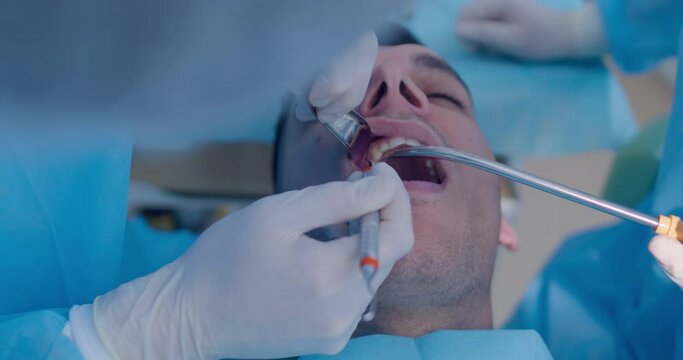 Close-up an Arabian male patient is having teeth cleaning procedure by the dentist who is using a dental probe and dental tool while an assistant is holding a saliva ejector.
