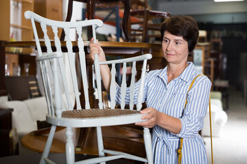 Fototapeta premium Mature female is choosing old armchair for home in antique shop.