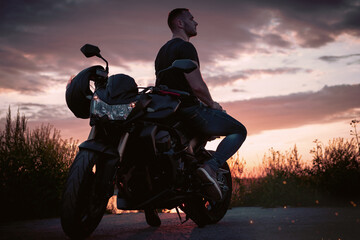 Obraz na płótnie Canvas Young handsome male biker, is sitting on a motorbike and enjoying the sunset near the shore.