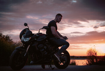 Young handsome male biker, is sitting on a motorbike during the sunset, thinking about life with his head down.