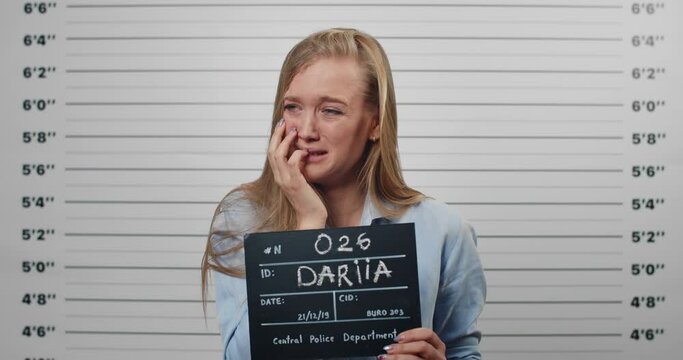 Mugshot of arrested young woman crying while holding sign for photo in police station. Millennial female person looking to camera while standing front of metric lineup wall.