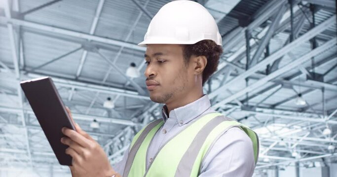 Close up of African American man architect standing alone in warehouse at work and texting on tablet using professional app Happy guy inspector in helmet typing on device, side view, building industry