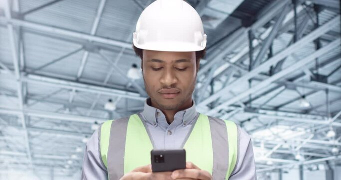 Close up portrait of young male African American engineer in helmet texting on smartphone searching social network app standing at construction site. Guy wearing safety equipment. Building industry