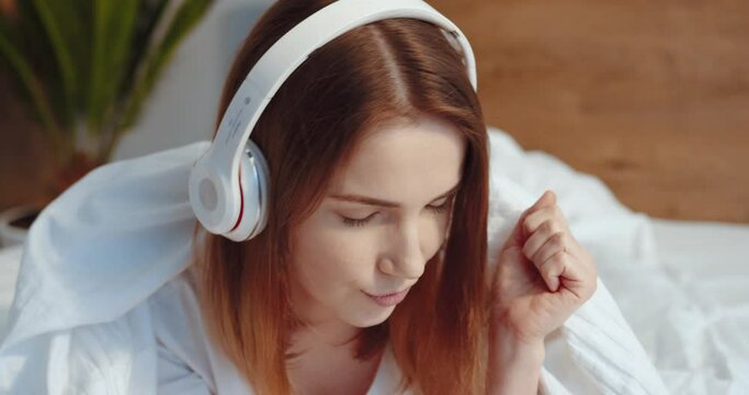 Horizontal view of the peaceful girl in modern wireless headphones relaxing at comfortable bed and listening to music. Happy calm young woman with ginger hair enjoy good quality sound. Stress free
