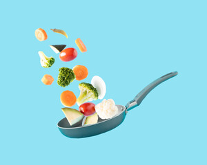 Chopped fresh vegetables fly over a black pan on a light blue background. Concept of flying food and healthy eating.
