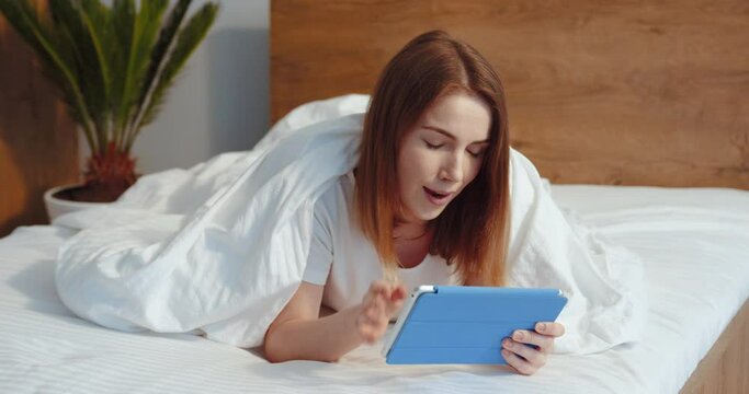 Horizontal view of the caucasian girl with ginger hair using her tablet while speaking with family at home. Lady laying at the stomach at the bed and waving to the camera at her cozy flat. Morning