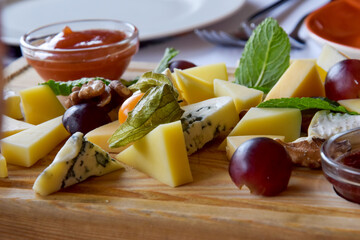 On a wooden plank a cheese platter made from several types of cheese and decorated with grapes. Dor Blue, Pecorino Romano, Tete de Moine, Maasdam, Parmesan, Camembert are delicious appetizers for wine