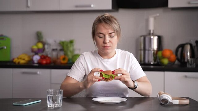 Health Conscious Female Taking One Last Bite Of Her Hamburger And Pushing Plate Away While Sitting At Kitchen Table. Adult Woman Deciding To Quit Eating Fast Food And Leading Healthy Lifestyle