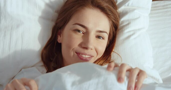 Don't want to get up. Top view of the coquettish ginger woman peeking up from the white blanket, looking at the camera with cute smile and hiding under the blanket at the bed