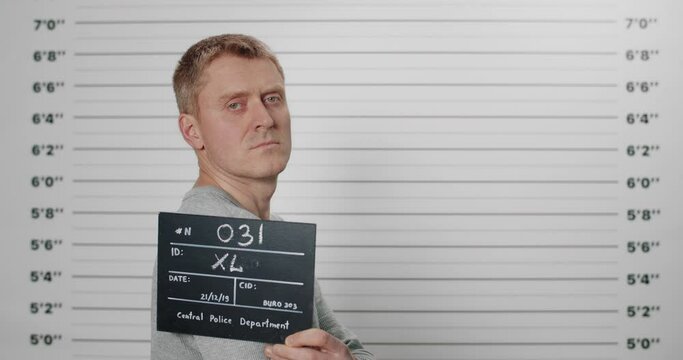 Side profile mugshot of man holding sign while being photographed in police department. Male criminal with green eyes turning head and looking to camera. Concept of crime.