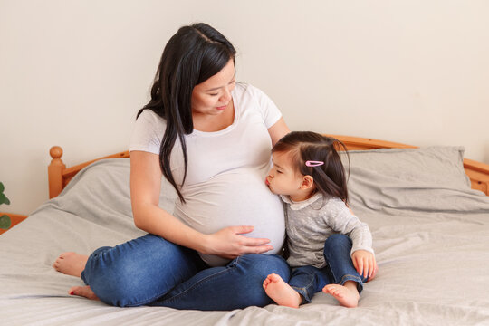 Asian Chinese Pregnant Woman With Toddler Girl Sitting On Bed At Home. Girl Daughter Kid Kissing Mom Belly. Mother And Baby Daughter Expecting Waiting For New Family Member. Ethnic Diversity.