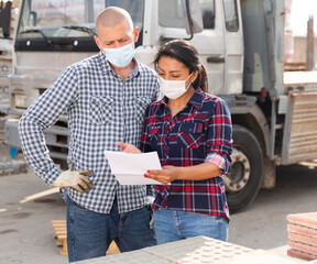 Manager in protective mask gives instructions to the car driver at the building materials storage...