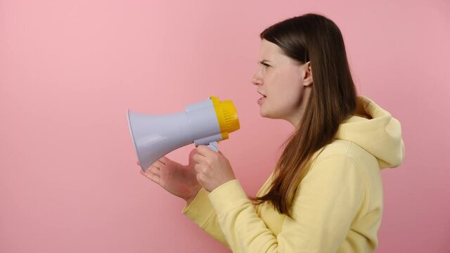 Side profile view sad protesting caucasian woman scream in megaphone, angry girl 20s does not hold back emotions, to shout furiously, wears yellow hoodie, posing isolated on pink color wall background