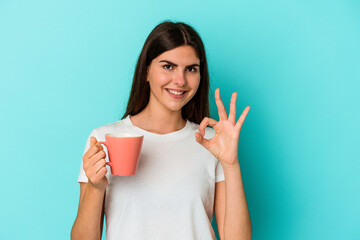 Young caucasian woman holding a mug isolated on blue background cheerful and confident showing ok...