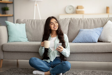 Fototapeta premium Online life. Gorgeous Indian woman sitting on floor with smartphone and cup of coffee, working or studying from home