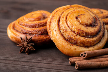 Cinnamon buns close up on dark wooden table. Anise star and cinnamon sticks near spiral rolls, aromatic condiments.