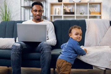 Afro american man in casual wear working on laptop and taking care of little baby boy. Young multitasking father with son at home. Concept of of distance work and parenting.