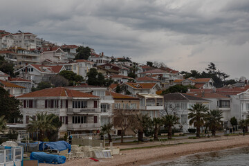 View from the sea on the island of Heybeliada in Istanbul. The slope shore is covered with many white summer cottages with brown tiled roofs. Beach with pebbles. Dramatic grey clouds in a stormy sky