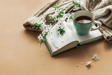 Obraz na płótnie Canvas Spring morning coffee. A cup of coffee, an open book and a warm sweater on the background of a bouquet of flowering branches. Cozy morning. Copy space. 
