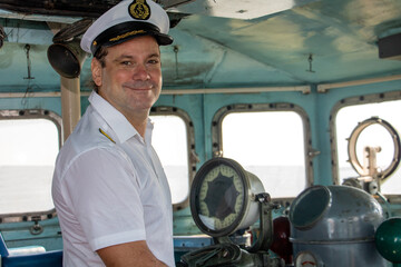 A captain standing in the wheelhouse of ship and looking to camera. - obrazy, fototapety, plakaty