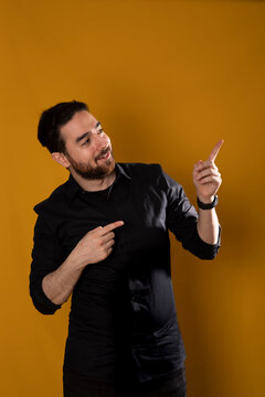 S&uuml;dl&auml;ndischer junger Mann steht vom gelben Hintergrund und zeigt mit den Fingern oben rlinks. In der Richtung schauet er auch nach. Er ist schwarz bekleidet und hat schwarzen Bart.  Studio Foto.