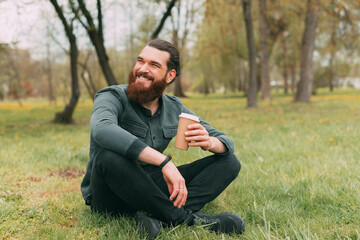 Naklejka na ściany i meble Portrait of smiling bearded man sitting in park on grass and drinking cup of coffee to go
